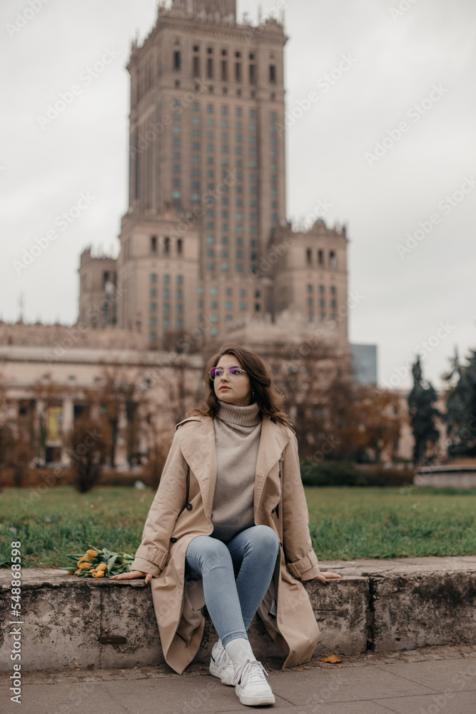 A walk near the Palace of Culture. A girl with yellow tulips against the background of the Palace of Culture in Poland. A pretty girl in glasses with flowers walks in Europe city.