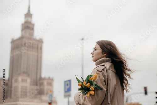A young girl in glasses with flowers against the background of the Palace of Culture in Warsaw. A pretty girl in glasses with flowers walks in Warsaw.
