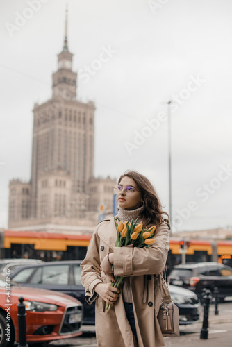 A young girl in glasses with flowers against the background of the Palace of Culture in Warsaw. A pretty girl in glasses with flowers walks in Warsaw.