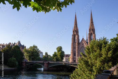 Church with two towers in France surrounded by a river, built with a reddish stone and near trees