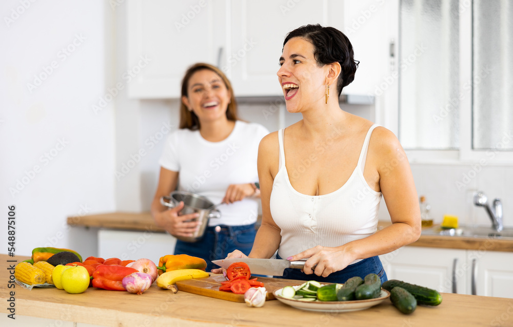 Happy young latin american girl standing in cozy kitchen interior at home with girlfriend, slicing fresh vegetables for salad for dinner. LGBT couple relationship concept..