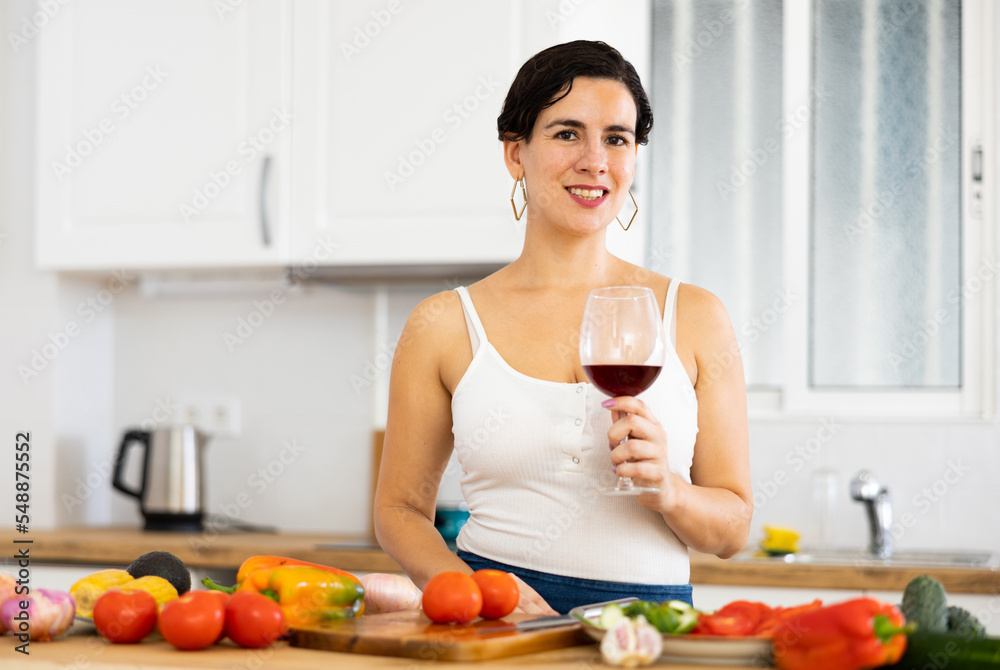 Cheerful young Hispanic woman relaxing after work at home, enjoying glass of red wine while preparing vegetable salad for dinner