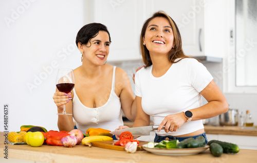 Two happy female friends preparing salad and drinking red wine together in modern kitchen