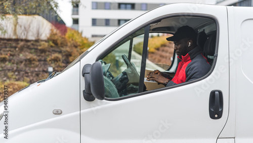 Young adult black delivery man in red pullover sitting in white van working on laptop computer. Delivery service. Horizontal outdoor shot. High quality photo