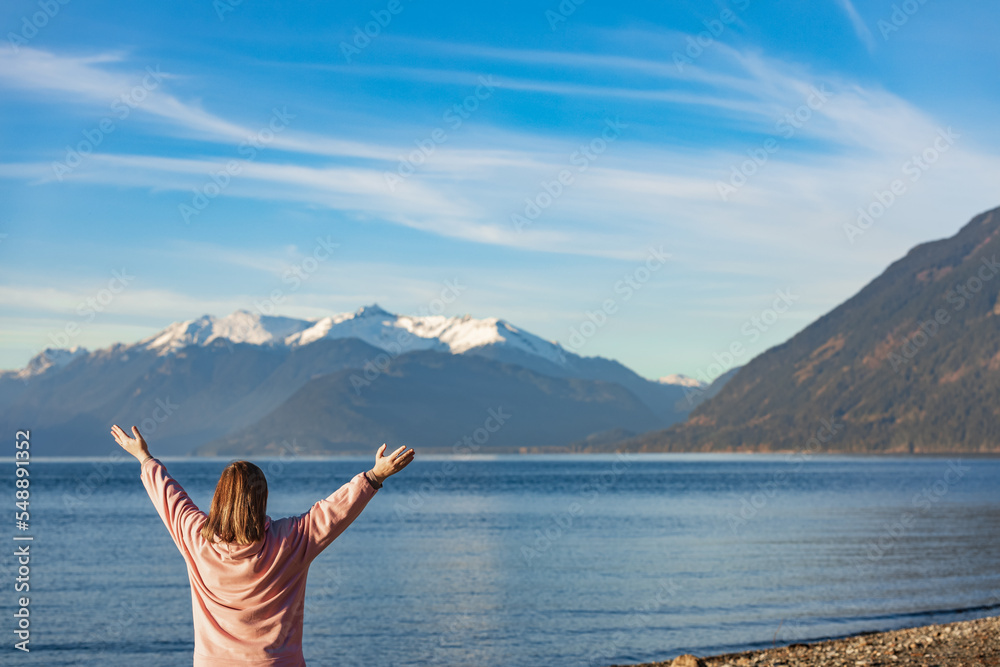 Happy woman with hands up standing by the lake and mountain in the distance. Woman enjoying the view of beautiful nature