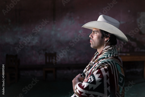 A young Mexican is sitting with crossed arms inside of a church wearing a hat and a poncho