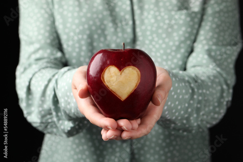 Fototapeta Naklejka Na Ścianę i Meble -  Woman holding red apple with carved heart on black background, closeup