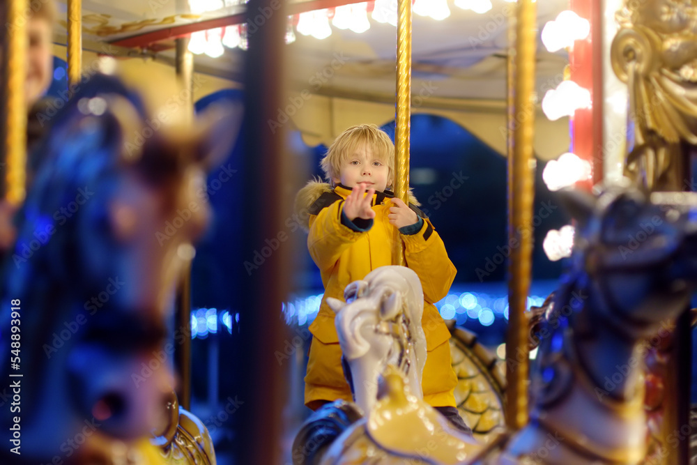 Cute blonde boy enjoying Christmas fair. Little child riding on a vintage carousel (merry go round). Outdoors entertainment activity for children on winter holidays