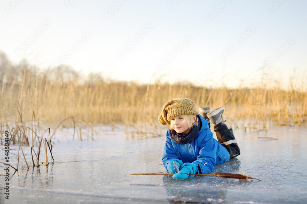 Cute preschool boy is playing on the ice of a frozen lake or river on a cold sunny winter sunset. Child having fun with icicle and dry reed plant during family hiking. Kids outdoor games - obrazy, fototapety, plakaty 