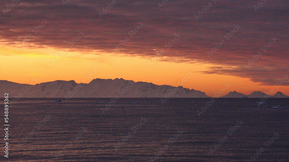 Pink clouds in an orange sky, with mountains and icebergs in silhouette, at Cierva Cove, Antarctica, at sunset