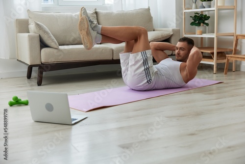 Man sports, watching tape of workout on phone and repeating exercises sports blogger with laptop training online, pumped up man fitness trainer works out at home, the concept of health and body beauty