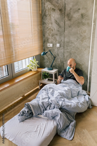 Man using smartphone and drinking coffee in bed © STUDIO TAURUS