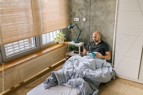 Man using smartphone and drinking coffee in bed