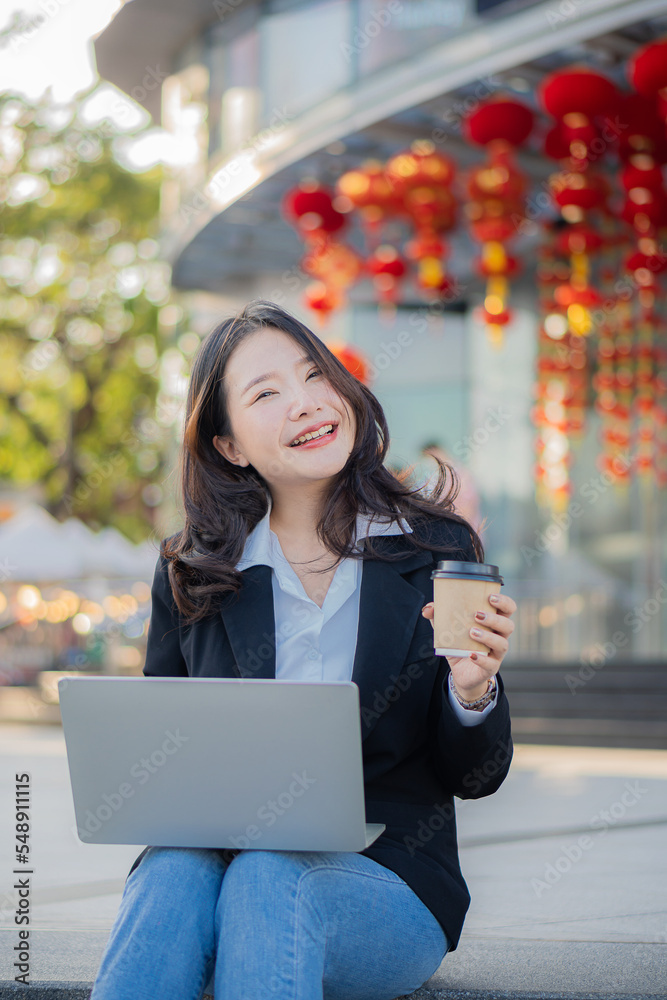 Fototapeta premium Asian businesswoman using the phone while having coffee while traveling is the new normal in the city, woman working with laptop outdoors.