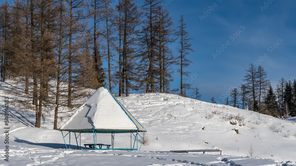 In the valley there is a gazebo with a layer of snow on a pyramidal roof. Bare trees on a hill against a blue sky. Altai