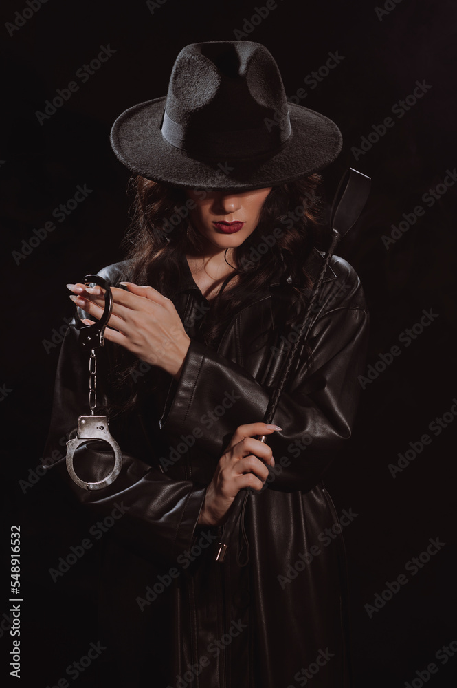 Fototapeta premium Sexy female dominant mistress with whip flogger and handcuffs for BDSM sex with submission and domination in a leather raincoat and hat in noir style