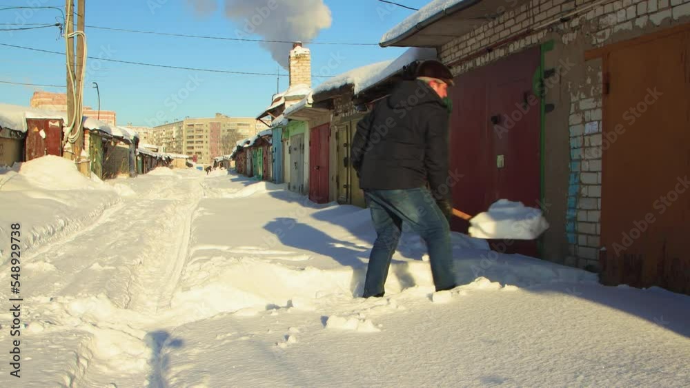 A cheerful man clears the passage to the garage from snow with a wooden shovel. Large snowdrifts around after a snowstorm, sunny day. City life, heavy snowfall. UHD 4K. A cheerful man clears the passage to the garage from snow with a wooden shovel. Large snowdrifts around after a snowstorm, sunny day. City life, heavy snowfall. UHD 4K.