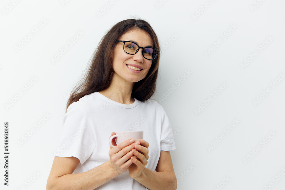 a sweet, beautiful, pleasant woman in glasses and a white t-shirt stands holding a cup in her hands and smiles sweetly.Horizontal photo on a light background a sweet, beautiful, pleasant woman in glasses and a white t-shirt stands holding a cup in her hands and smiles sweetly.Horizontal photo on a light background