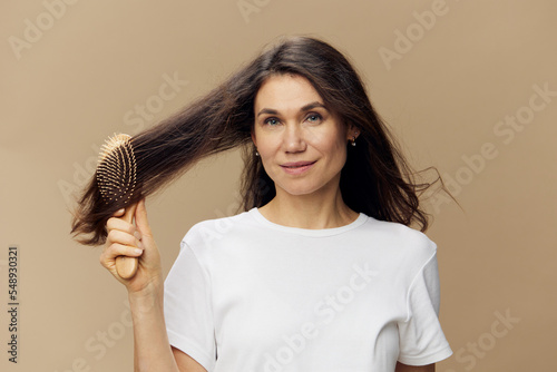 a sweet, beautiful middle-aged brunette woman stands pleasantly smiling on a beige background and combs her long hair with a wooden massage comb. Horizontal photo on
