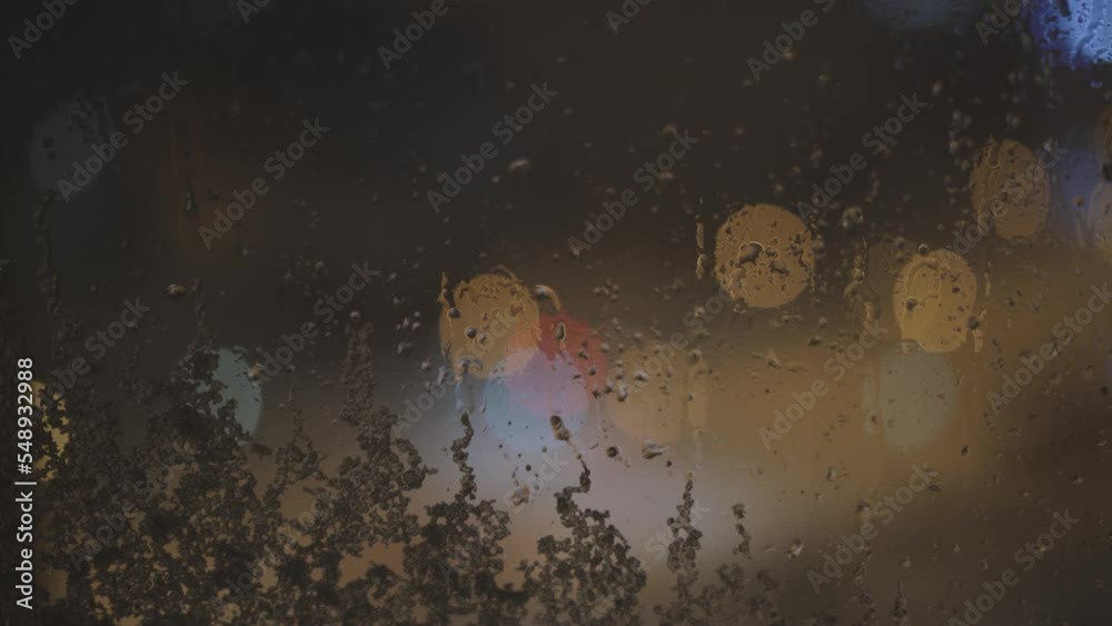 Snow-covered window in snowstorm at night, close-up. Snow falls on glass, melts and flows down. Abstact, blurred round multi-colored city lights outside the window. Snow-covered window in snowstorm at night, close-up. Snow falls on glass, melts and flows down. Abstact, blurred round multi-colored city lights outside the window.