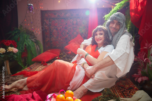 Portrait of young arabian muslim couple in traditional clothes in cozy red room. Fhoto shoot in easten style with male and female model like in a harem with a sultan and an odalisque photo