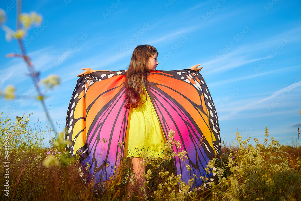Beautiful little girl in yellow dress and in colorful bright butterfly wings in the field with green grass, yellow flowers and blue sky in a sunny summer or spring day