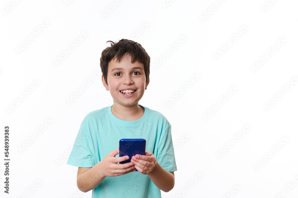 Waist-length portrait, isolated over white background of a smiling happy friendly teenage boy in blue turquoise t-shirt, using mobile cell phone, browsing surfing internet. People, lifestyle concept Waist-length portrait, isolated over white background of a smiling happy friendly teenage boy in blue turquoise t-shirt, using mobile cell phone, browsing surfing internet. People, lifestyle concept