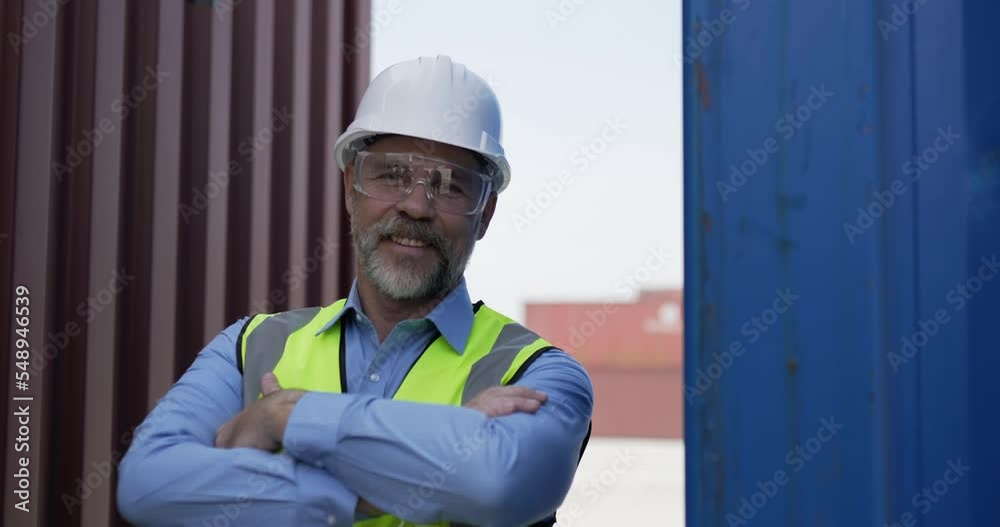 Handheld shot, Portrait of middle-aged Caucasian Business engineer man wearing helmet stand with arms crossed, turn to smile and look at the camera while working in container terminal port Handheld shot, Portrait of middle-aged Caucasian Business engineer man wearing helmet stand with arms crossed, turn to smile and look at the camera while working in container terminal port
