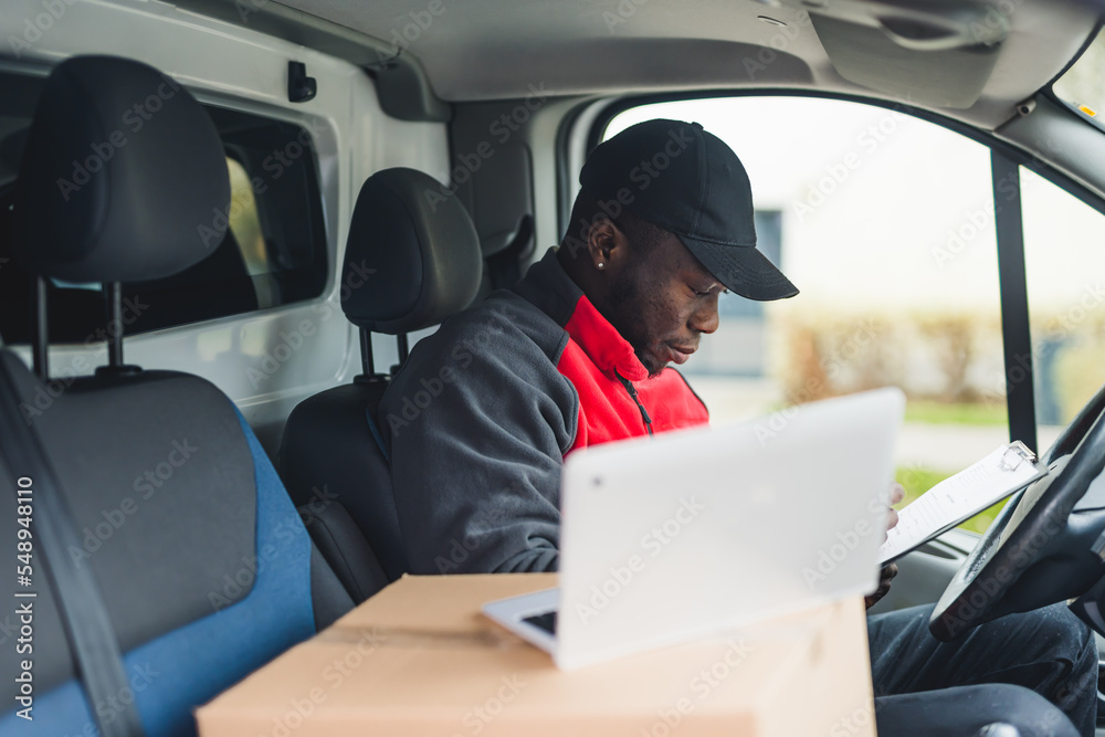 Young adult black delivery man sitting in driver seat of van looking through documents with open laptop computer on the side. Horizontal shot. High quality photo