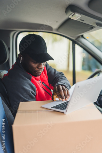 Young adult black delivery guy wearing work uniform sitting in driver seat of van working on laptop computer on top of package. Vertical shot. High quality photo