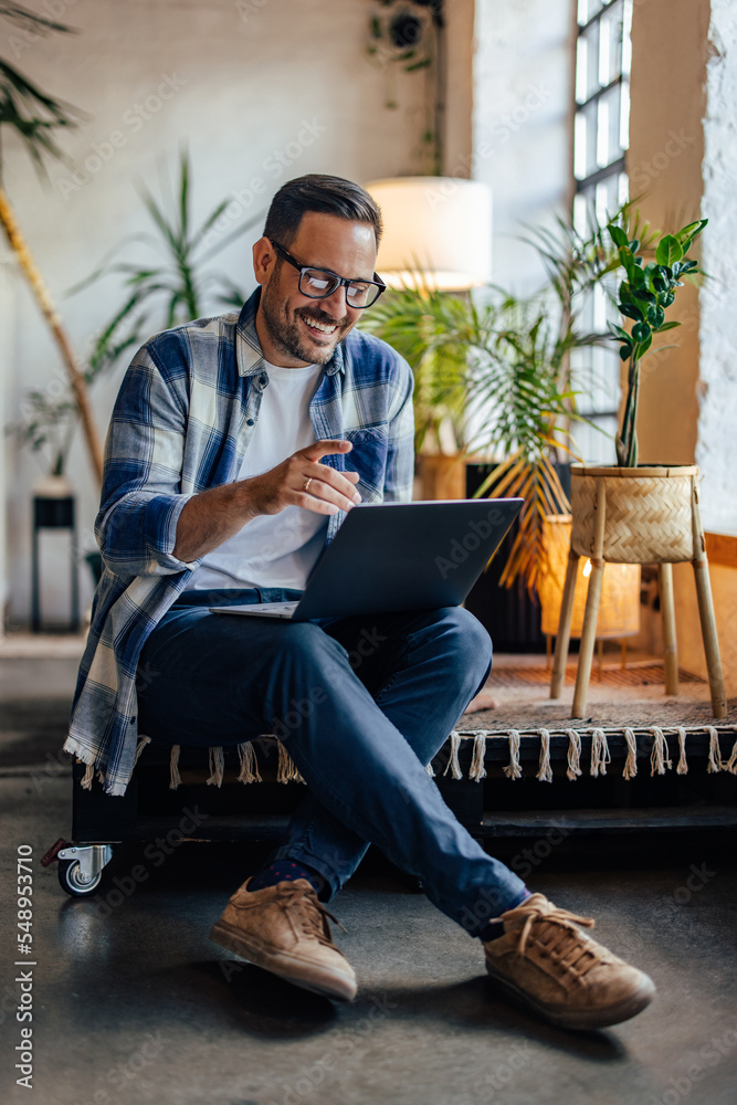 Smiling businessman sitting somewhere in the co-working space, talking with someone, over the laptop.