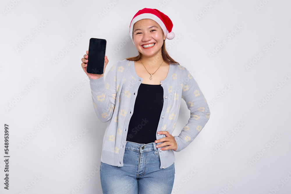 Portrait of a smiling beautiful young Asian woman in a Christmas hat isolated on white background. smiling girl standing and showing a blank smartphone screen Portrait of a smiling beautiful young Asian woman in a Christmas hat isolated on white background. smiling girl standing and showing a blank smartphone screen