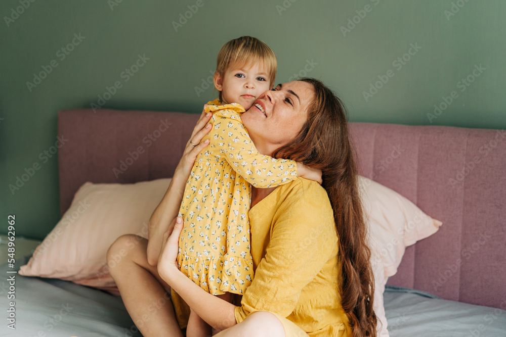 Red-haired mother hugs her little toddler daughter while sitting in bed. Red-haired mother hugs her little toddler daughter while sitting in bed.