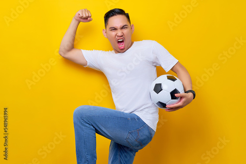 Excited young Asian man 20s wearing a white t-shirt supporting a football sports team, holding in hand a soccer ball and watching tv live stream do winner gesture isolated on yellow background © Bangun Stock Photo