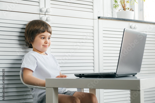 5 year boy sit at the table uses laptop and looks at the screen. Child is doing homework lesson, playing video games, studying IT development, watching cartoon. Home distance online education concept