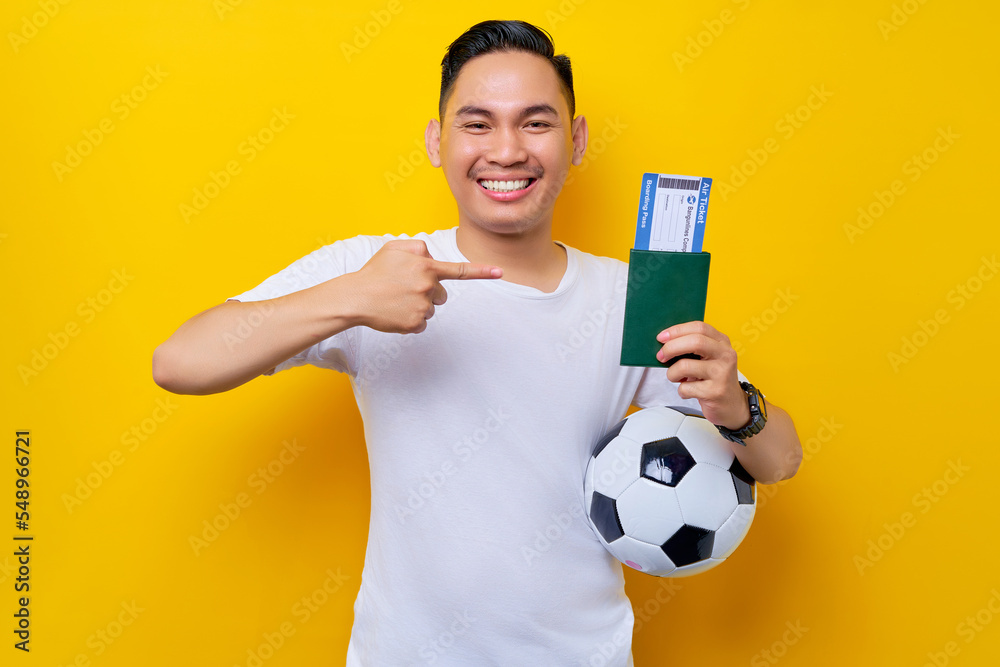 excited young Asian man football fan wearing a white t-shirt carrying a soccer ball and pointing a finger at a passport ticket boarding pass isolated on yellow background excited young Asian man football fan wearing a white t-shirt carrying a soccer ball and pointing a finger at a passport ticket boarding pass isolated on yellow background