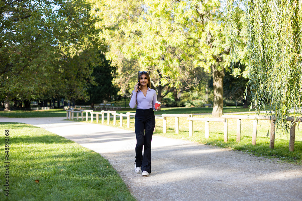 Woman walks in the park and has a phone call with her smartphone. She has brown hair, long sleeve, holds a coffee mug in the hand. Outdoor, public park, relaxed position and summer. Woman walks in the park and has a phone call with her smartphone. She has brown hair, long sleeve, holds a coffee mug in the hand. Outdoor, public park, relaxed position and summer.