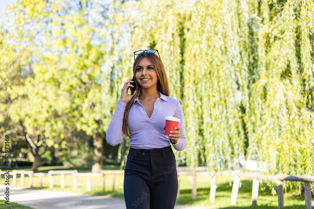 Woman walks in the park with smartphone. She has brown hair, wearing a long sleeve and holds a coffee cup while she has a phone call. Outdoor, public park, relaxed position and summer. Woman walks in the park with smartphone. She has brown hair, wearing a long sleeve and holds a coffee cup while she has a phone call. Outdoor, public park, relaxed position and summer.