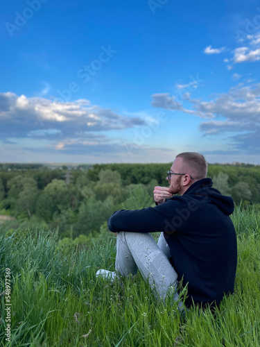 Rear view of thoughtful young man sitting on grass against blue sky. © Vitalina