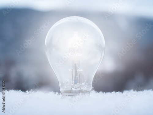 Close-up of a lit light bulb sticking up from soft snow with a blurred winter landscape in the background