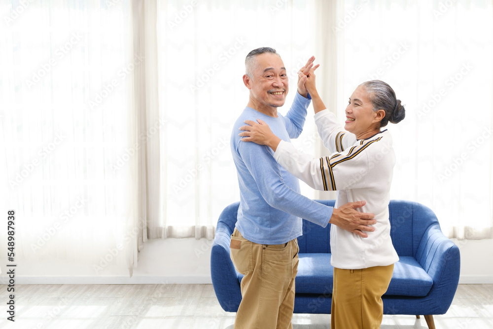Romantic and cheerful Asian well-being senior couple enjoy dancing and holding hands to music together with smiles and happiness in the living room. Senior retirement activity at home. Romantic and cheerful Asian well-being senior couple enjoy dancing and holding hands to music together with smiles and happiness in the living room. Senior retirement activity at home.