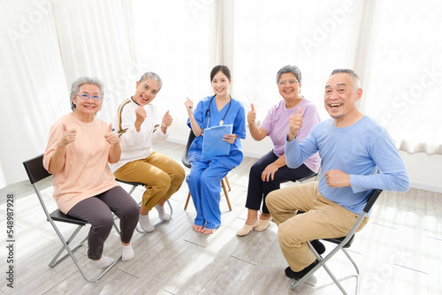 The caregiver therapist sits with a group of Asian senior people with thumbs up in a circle for checking physical and mental health in a group elderly therapy session. The nursing home facilitates