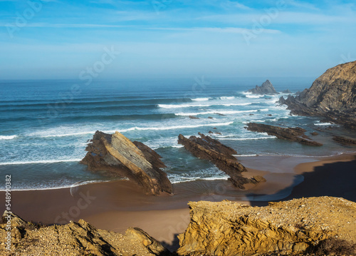 View of empty Alteirinhos sand beach with ocean waves, cliffs, stones and rocks in morning golden light at wild Rota Vicentina coast, Odemira, Portugal. photo