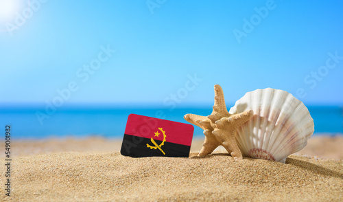 Tropical beach with seashells and Angola flag. The concept of a paradise vacation on the beaches of Angola.
