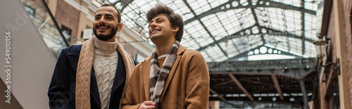 young and fashionable gay men smiling and looking away under transparent roof outdoors, banner.