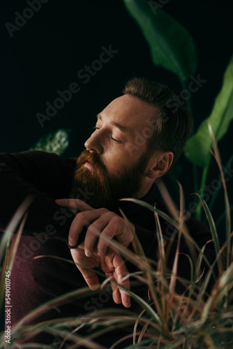 Fashionable man in jumper looking away near blurred plants isolated on black. © LIGHTFIELD STUDIOS