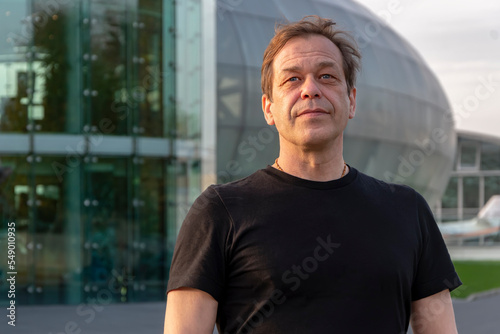 Street portrait of a man 40-50 years old in a black T-shirt on a blurred background of a glass building. Perhaps he is a tourist or a traveler, an actor or a truck driver, a loader or a pensioner,