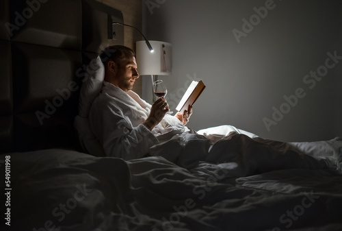 Midle-aged man relaxing rin bed reading book holding a glass of red wine with bedside lamp turned on. Evening relaxation, hobbies, free time concept. Adulthood concept. photo