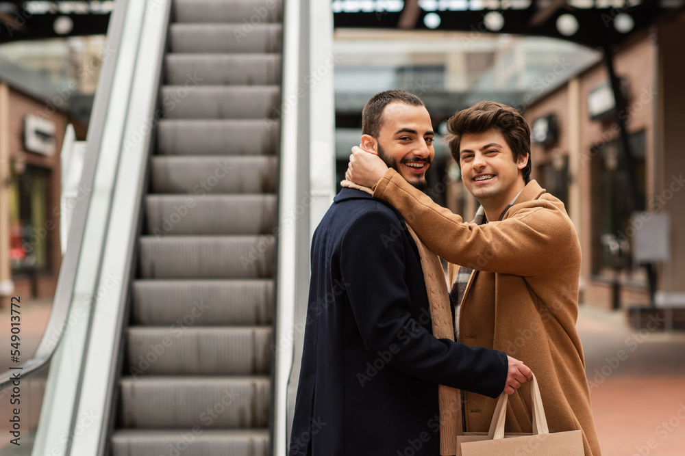 happy gay man in beige coat hugging neck of bearded boyfriend holding shopping bags near escalator. happy gay man in beige coat hugging neck of bearded boyfriend holding shopping bags near escalator.