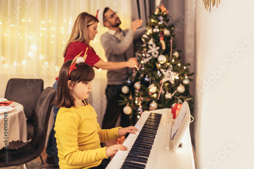 Young parent decorate the Christmas tree enjoying music that their daughter playing on piano during Christmastime © Mediteraneo